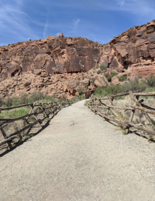 A dirt path leads through a rocky landscape, flanked by wooden fences and surrounded by red rock formations.