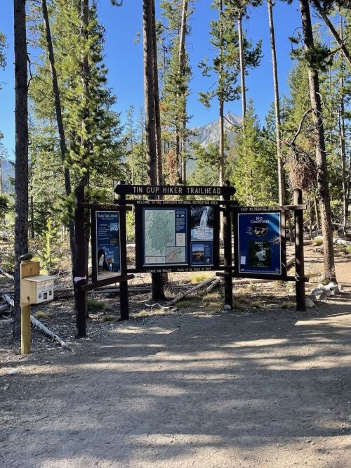 Trailhead sign for Tin Cup Hiker, surrounded by trees and mountains, with maps and information displayed.