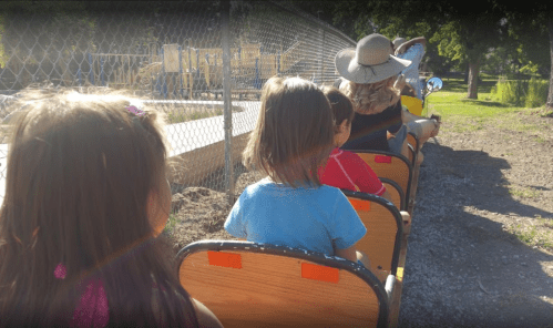 Children sitting on a small train ride, enjoying a sunny day at a park.