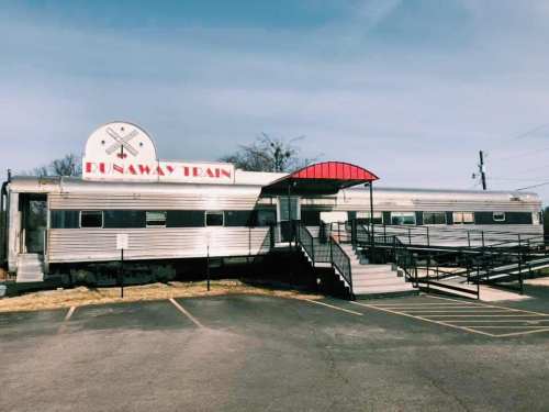 A vintage silver train car named "Runaway Train" with a red awning, parked beside a paved area and blue sky.
