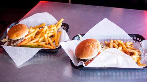 Two trays with burgers and crispy fries, served on white paper, on a metallic surface.