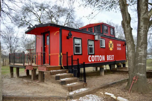 A bright red caboose labeled "COTTON BELT" sits on a raised platform surrounded by trees.