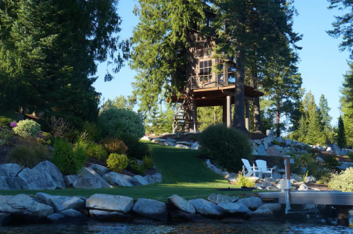 A treehouse perched on tall stilts surrounded by lush greenery and rocks, with a dock and chairs by the water.