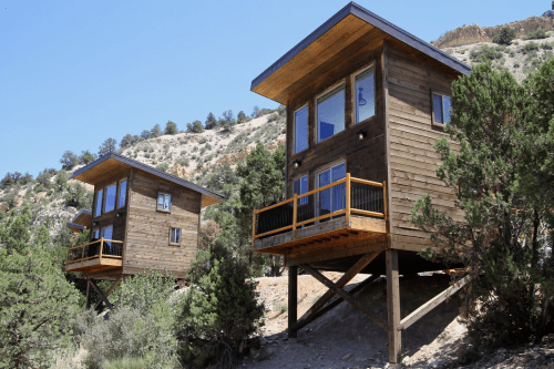 Two modern wooden cabins on stilts, surrounded by greenery and set against a rocky hillside under a clear blue sky.
