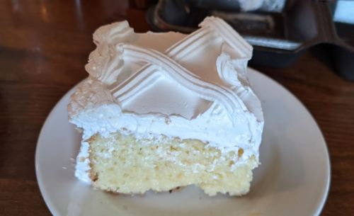 A slice of yellow cake topped with white frosting on a plate, set on a wooden table.