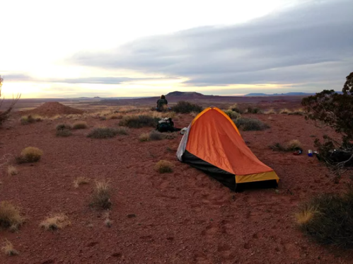 A tent set up on red desert terrain with sparse vegetation, under a cloudy sky at sunset.