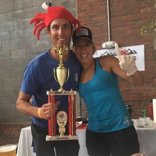 Two smiling individuals hold a trophy, one wearing a crab hat, celebrating a lobster roll competition.