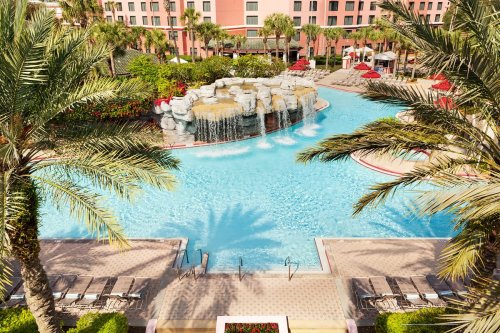 A resort pool with a waterfall, surrounded by palm trees and lounge chairs, set against a pink hotel backdrop.
