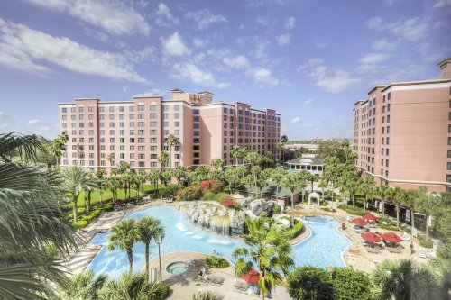 A resort with a large pool surrounded by palm trees and colorful landscaping, featuring a pink hotel in the background.
