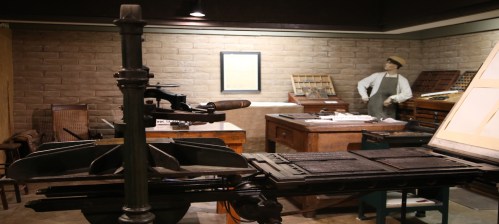 A vintage printing press in a historical workshop, with wooden tables and a mannequin dressed in period attire.