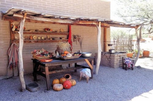 A rustic outdoor kitchen with a wooden table, pots, pumpkins, and decorative items under a shaded structure.