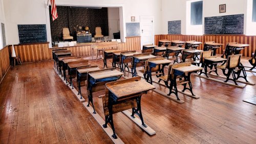 A vintage classroom with wooden desks, a blackboard, and a stage at the front, featuring two chairs and an American flag.