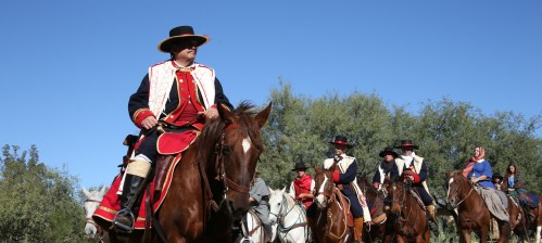 A group of horse riders in traditional attire, riding through a sunny landscape with trees in the background.