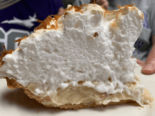A slice of coconut cream pie with a fluffy meringue topping and a golden crust, displayed on a plate.