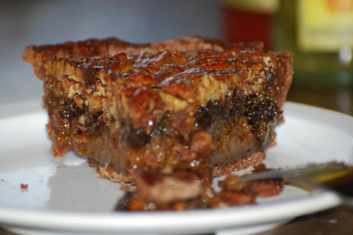 A slice of pecan pie on a white plate, showcasing a rich filling and a golden-brown crust.