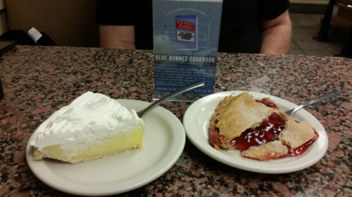 A slice of lemon pie with whipped cream and a serving of cherry cobbler on a table with a menu in the background.