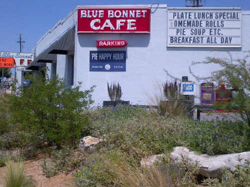 Signage for Blue Bonnet Cafe featuring "Pie Happy Hour" and daily specials, with a desert landscape in the foreground.