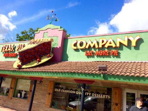 Colorful storefront of "Texas Pie Company" with a large pie sign and the slogan "Eat More Pie." Bright blue sky above.