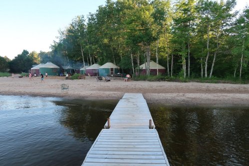 A wooden dock leads to a sandy beach with yurts and trees in the background, with people enjoying the outdoor space.