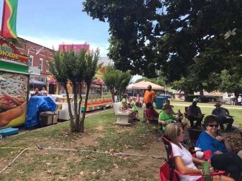 A lively outdoor scene with people sitting on chairs, food stalls, and trees in a park setting.