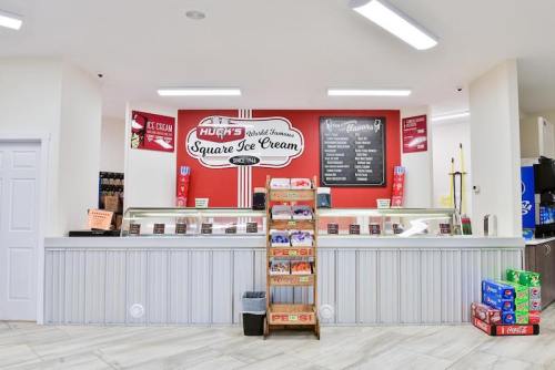 A bright ice cream shop counter with a menu, ice cream tubs, and a display of snacks and drinks.