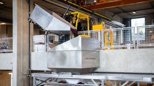 A stainless steel machine for processing materials, with a large hopper and conveyor system in an industrial setting.