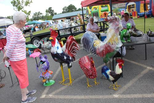 A woman admires colorful metal rooster sculptures at an outdoor market with various stalls in the background.