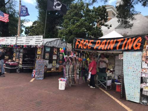 A vibrant outdoor market scene featuring a face painting booth and various vendor stalls with colorful merchandise.
