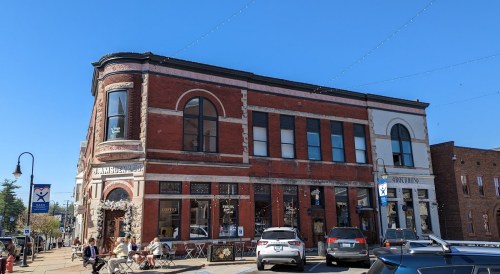 Historic brick building with large windows, outdoor seating, and cars parked nearby under a clear blue sky.