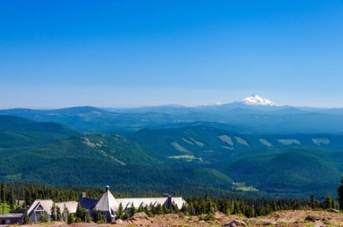 A panoramic view of mountains and forests under a clear blue sky, with a distant snow-capped peak visible.