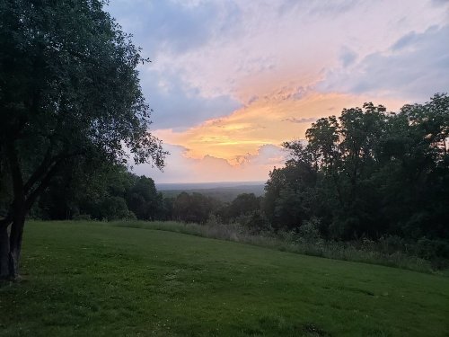 A serene landscape at sunset, featuring a grassy field, trees, and a colorful sky with clouds.