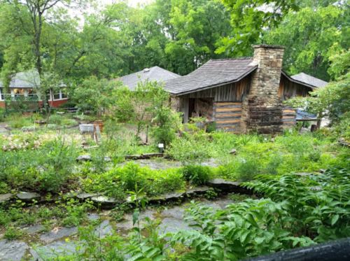 A lush garden surrounds rustic log cabins, with greenery and trees creating a serene, natural setting.
