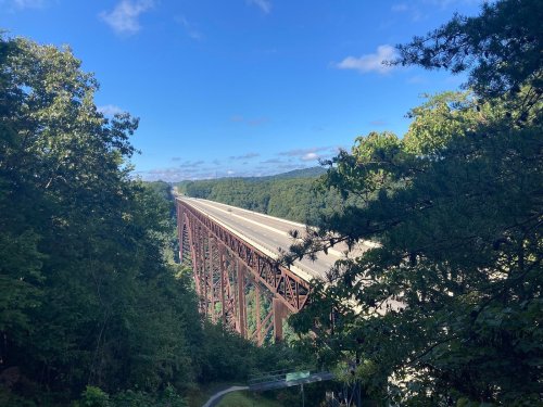A tall, rust-colored bridge spans a valley, surrounded by lush green trees under a clear blue sky.