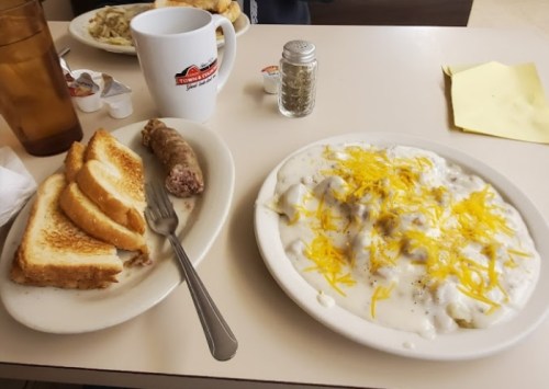 A plate of biscuits and gravy topped with cheese, alongside toast and a sausage link on a table with a coffee mug.
