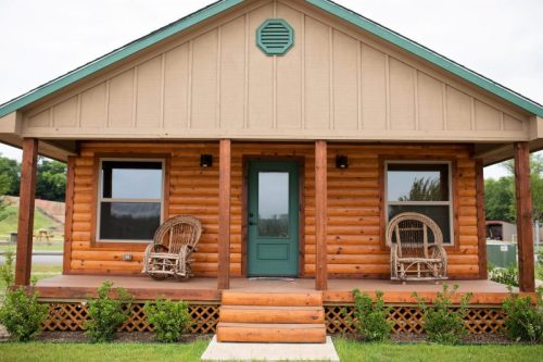 A cozy log cabin with a green door, two rocking chairs on the porch, and a grassy area in front.