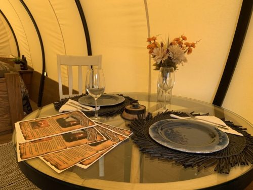A cozy dining setup inside a tent, featuring a glass table, plates, menus, and a vase of flowers.