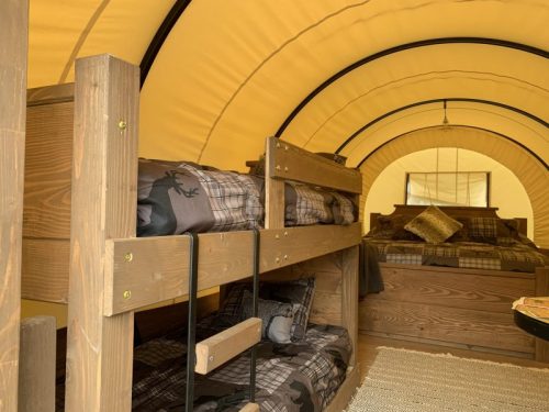 Interior of a cozy tent with wooden bunk beds and a double bed, all featuring plaid bedding and a warm yellow canopy.