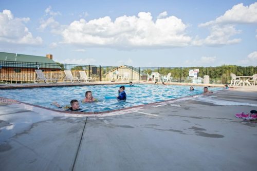Children swimming in a pool on a sunny day, with lounge chairs and a wooden building in the background.