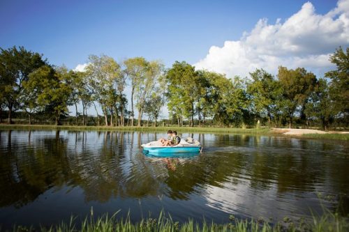 A small blue paddle boat with two people floats on a calm pond surrounded by trees under a clear blue sky.