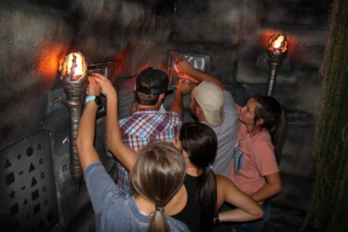 A group of five people working together to solve a puzzle in a dimly lit, themed escape room.