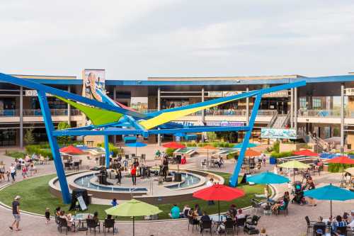 A vibrant outdoor plaza with colorful umbrellas, a fountain, and people enjoying the space around shops and restaurants.