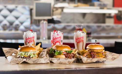 Three burgers with toppings and fries, accompanied by milkshakes topped with cherries, on a table at a diner.