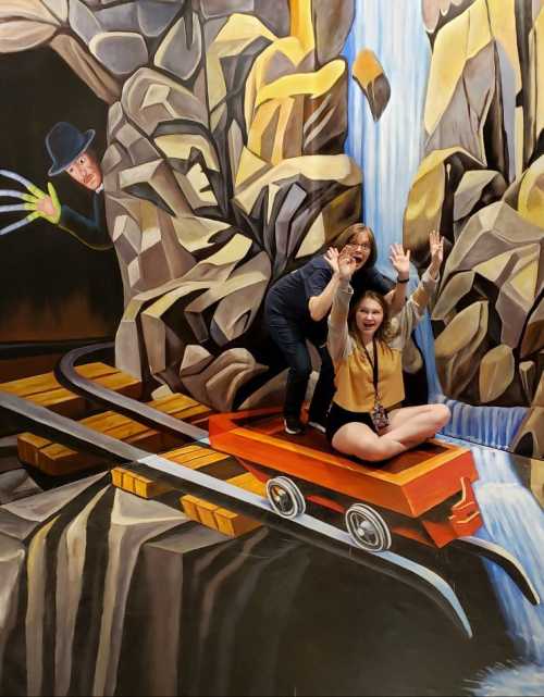 Two women pose on a 3D mural of a mine cart near a waterfall, with one playfully raising her hands in excitement.