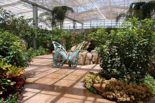 A vibrant indoor garden with lush plants, a butterfly sculpture, and a small water feature under a glass roof.