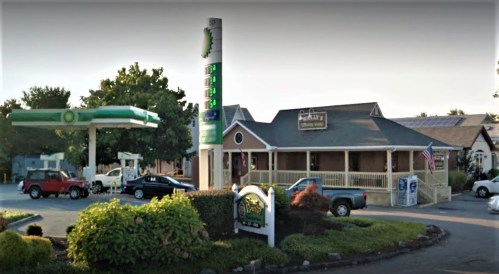 A gas station and restaurant with a sign, surrounded by trees and parked cars in a sunny setting.