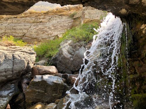 A waterfall cascades over rocks, framed by a rocky overhang and surrounded by greenery.
