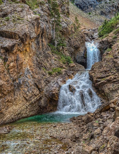 A scenic waterfall cascades down rocky cliffs into a clear pool, surrounded by lush greenery and rugged terrain.