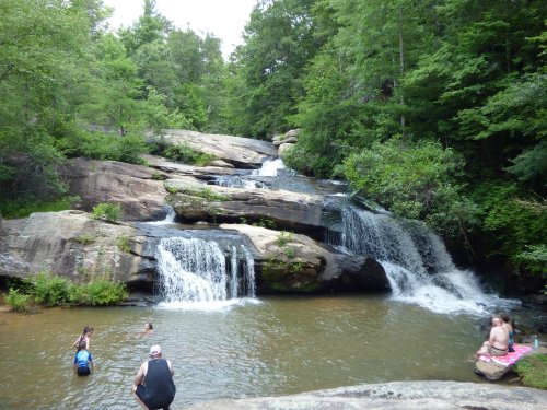 A serene waterfall cascades over rocks into a swimming hole, surrounded by lush greenery and people enjoying the water.