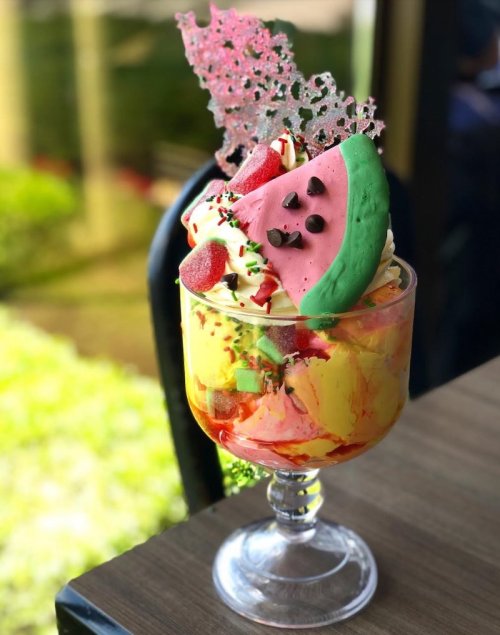 A colorful dessert in a glass, featuring layers of ice cream, fruit, and decorative toppings like chocolate and a green cookie.