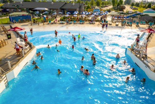 A busy water park scene with people swimming in a wave pool, surrounded by lounge chairs and umbrellas.